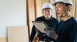 © 望見 柿元 - 日本人の建設業シニア男性が、現場作業でDX化されたタブレット端末を使い若手男性に育成指導を行う様子。屋内の建築現場で打ち合わせをするネイビーの作業服を着た二人、ヘルメット着用の人物。