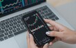 © joeycheung - Person analyzes stock charts on phone with laptop in background for