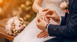 © Esther - Groom placing wedding ring on bride's finger during an outdoor ceremony, with a bridal bouquet nearby.