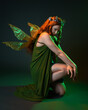 © faestock - portrait of female model wearing fantasy fairy costume, green dress, cloak, butterfly wings and crown. full length sitting pose, kneeling on floor, dark studio background.