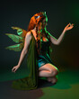 © faestock - portrait of female model wearing fantasy fairy costume, green dress, cloak, butterfly wings and crown. full length sitting pose, kneeling on floor, dark studio background.