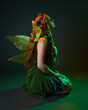 © faestock - portrait of female model wearing fantasy fairy costume, green dress, cloak, butterfly wings and crown. full length sitting pose, kneeling on floor, dark studio background.