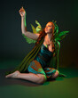 © faestock - portrait of female model wearing fantasy fairy costume, green dress, cloak, butterfly wings and crown. full length sitting pose, kneeling on floor, dark studio background.