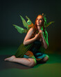 © faestock - portrait of female model wearing fantasy fairy costume, green dress, cloak, butterfly wings and crown. full length sitting pose, kneeling on floor, dark studio background.