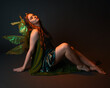 © faestock - portrait of female model wearing fantasy fairy costume, green dress, cloak, butterfly wings and crown. full length sitting pose, kneeling on floor, dark studio background.