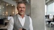 © nyul - Confident, successful middle aged businessman in office. Portrait of happy mid adult man smiling. Mature entrepreneur standing in corporate workspace. Senior male in business vest and white shirt.
