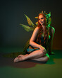 © faestock - portrait of female model wearing fantasy fairy costume, green dress, cloak, butterfly wings and crown. full length sitting pose, kneeling on floor, dark studio background.