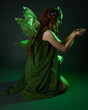 © faestock - portrait of female model wearing fantasy fairy costume, green dress, cloak, butterfly wings and crown. full length sitting pose, kneeling on floor, dark studio background.