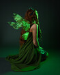 © faestock - portrait of female model wearing fantasy fairy costume, green dress, cloak, butterfly wings and crown. full length sitting pose, kneeling on floor, dark studio background.