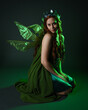 © faestock - portrait of female model wearing fantasy fairy costume, green dress, cloak, butterfly wings and crown. full length sitting pose, kneeling on floor, dark studio background.