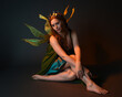 © faestock - portrait of female model wearing fantasy fairy costume, green dress, cloak, butterfly wings and crown. full length sitting pose, kneeling on floor, dark studio background.