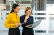 © Liubomir - Diverse businesswomen standing together. Smiling and discussing project data on a laptop and smartphone. Showcasing teamwork and modern corporate communication within a professional office environment