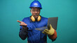 © Krakenimages.com - Man construction worker in blue hardhat and gloves holds laptop with open palm gesture toward camera in studio; confident problem solving.