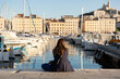 © Simplementbeau. - Femme vue de dos avec cheveux longs bruns assise sur le quai du Vieux Port de Marseille, regardant les bateaux et la colline avec la basilique Notre Dame de la Garde, lumière douce et chaude
