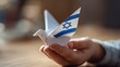 © Tina - A child's hand holds a paper dove with the Israeli flag design. The background is softly blurred, emphasizing the origami bird.