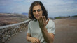 © Krakenimages.com - Man with long hair and glasses beckons with open hand beside a stone wall on a street outdoors; invitation connection.