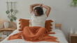 © Krakenimages.com - Young indian man on bed stretches arms up, yawning with orange blanket, tassel pillow and wicker bedside table in studio; morning serenity.