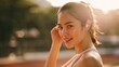 © tonpreecha - Young woman smiling while adjusting wireless earbuds in golden hour sunlight