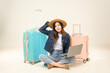 © waragron - A young woman in casual attire sits cross-legged with a laptop, flanked by pastel-colored suitcases, ready for travel.