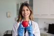 © fizkes - Head shot portrait of smiling Hispanic woman cardiologist dressed in white medical coat holding red heart object in her hands, posing for camera in clinic office. Heart health awareness, cardiology
