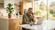 © SheulyAkter - A middle-aged man sitting at a kitchen table working on his laptop while talking on the phone