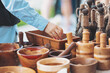 © Cristina - A person reaches into a wooden bowl at a craft fair. Wooden bowls, utensils, and other items are displayed on a table. People walk in the background under the sunlight