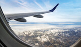 A view from an airplane window shows a silver wing soaring over a landscape of fluffy white clouds, dark blue water, and a coastal town.