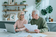 © AnnaStills - Senior Caucasian couple sitting at kitchen table, reviewing household bills on laptop and discussing finances at home