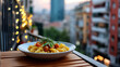 © Anastasiia Havelia - Close-up of a plate with fresh pasta and herbs on a tiny wooden balcony table, warm light from a nearby string of fairy lights, blurred view of dense urban architecture and sunset