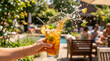 © Argiduna - Una mano sostiene un vaso de cóctel de frutas veraniego con un espectacular salpicón de agua en un ambiente de playa al aire libre. La bebida naranja contiene lima, bayas y menta sobre una mesa rústic