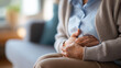 © Laura - Close-up of elderly person's hands clutching their stomach in apparent pain while sitting on a couch in a blurred living room background. Defocused living room. Elderly hands