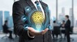 © Sonthana - A businessman in a suit holding a glowing digital light bulb with gears and data icons, symbolizing innovation and technology in a modern office setting.