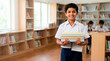 © Xenia - Smiling Indian schoolboy holding a stack of books in a library. Happy male student in uniform standing near bookshelves