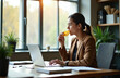 © miss irine - Businesswoman eats chips while typing on laptop at desk. She works in modern office, takes short snack break near window with plants. Professional woman uses computer for work.