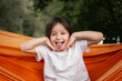 © Ananass - Young girl laughs with tongue out in bright orange hammock surrounded by green trees outdoors