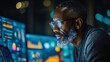 © Agung - Man of color with short hair and beard wearing glasses, intently focused on multiple glowing computer screens displaying data and charts in a dark room at night