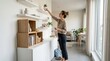 © Matweymax - Woman arranging a small potted plant on a wall shelf in a bright modern living room, with minimalist furniture and natural light, creating a calm home decorating scene for interior design themes