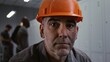 © Framestock - Close Up of Seasoned Factory Worker in Grey Work Jacket and Orange Helmet, Sitting on Bench and Looking Into Frame With Metal Lockers and Colleagues in Background. Industrial Preparation. Portrait.