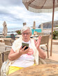 © luciano - Senior Woman Using Smartphone at Seaside Cafe Reacting with Surprise During Relaxing Coastal Vacation