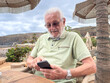 © luciano - Elderly Man Using Smartphone Outdoors At Beach Café During Relaxed Seaside Vacation