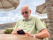 © luciano - Senior Man Using Smartphone at Beach Cafe Under Straw Umbrellas, Relaxing Outdoors on Holiday