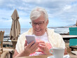© luciano - Senior Woman Smiling at Smartphone While Enjoying Coffee at Seaside Cafe on Relaxing Vacation