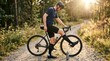© ЮРИЙ ПОЗДНИКОВ - Male cyclist in black gear adjusts bike on gravel path surrounded by trees and sunlight