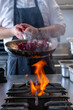 © Thomas - Scène dynamique d'un chef maniant une poêle au-dessus d'une flamme vive dans une cuisine professionnelle, faisant sauter des ingrédients colorés dans une vapeur légère.