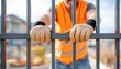 © dreamer - A worker in an orange vest grips metal bars, symbolizing prison or confinement, with a blurred background of a construction site or urban area.
