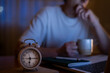 © Nanci - Young person yawning and drinking coffee in the early morning at home, experiencing insomnia due to late night remote work, prioritizing productivity with an alarm clock suggesting an early start