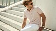 © watcharee - A stylish man wearing sunglasses and a light pink polo shirt sits on outdoor steps, looking to the side in a relaxed, casual pose.