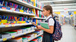 © Natali - A child stands in a store aisle looking at school supplies. She holds notebooks and a binder while wearing a backpack. The shelves are filled with colorful items