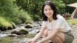 © AiPhoto - キャンプ場の川辺でしゃがんで川の水に触れる笑顔の女性