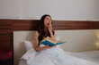 © Parichat - Young woman yawning reading book in bed